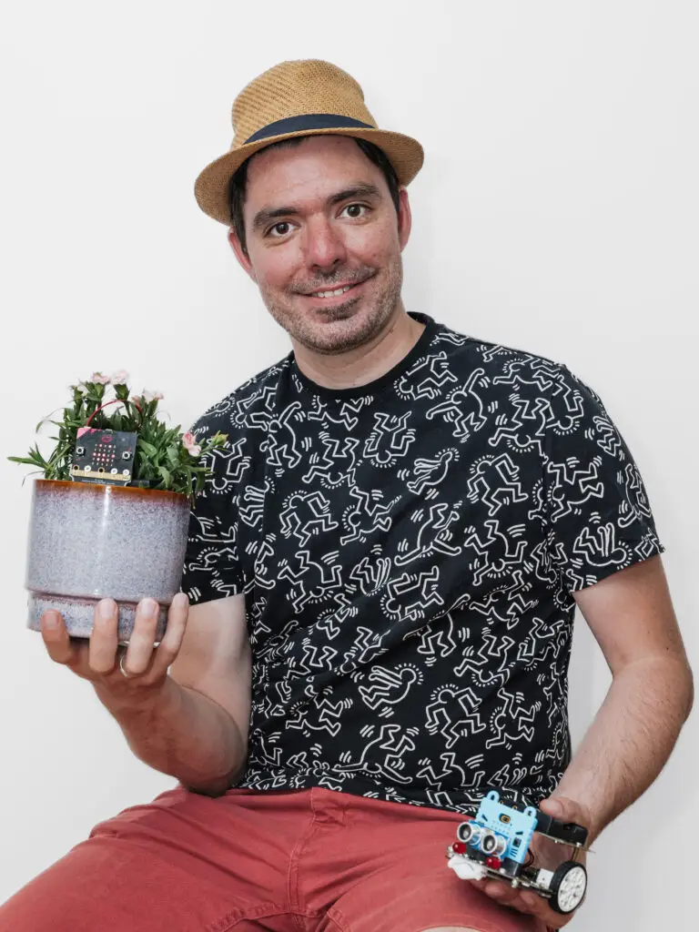 Tommy Berben, souriant et coiffé d'un chapeau de paille, tient d'une main un pot de fleurs équipé d'une carte micro:bit et de l'autre un petit robot à roues.